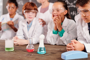 Three glass tubes with chemical compounds standing on teacher desk in chemical laboratory while schoolkids watching different reactions