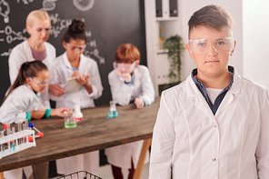 Contemporary pupil of secondary school in whitecoat and protective eyeglasses standing against classmates and teacher during lab work