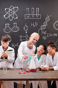 Group of clever students of secondary school in whitecoats and eyeglasses looking at their teacher showing chemical experiment by blackboard