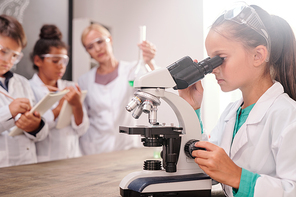 Clever serious schoolgirl in whitecoat looking in microscope by desk against her classmates and teacher making chemical experiment