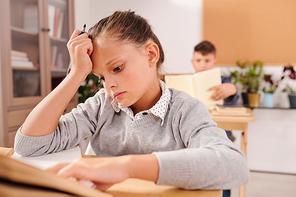 Serious cute schoolgirl looking at question in notebook and thinking of answer while sitting by desk at lesson and working individually