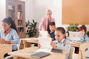 Contemporary schoolgirl and her classmates with touchpads sitting by desks and working individually while teacher standing by one of them