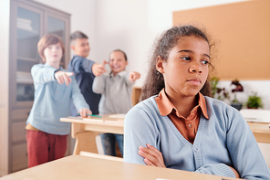 Offended mixed-race schoolgirl sitting by desk and crossing arms by chest on background of classmates bullying her at break in classroom
