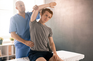 Mature physiotherapist in uniform supporting arm of young patient sitting on medical couch during sidebend exercise in rehabilitation clinics