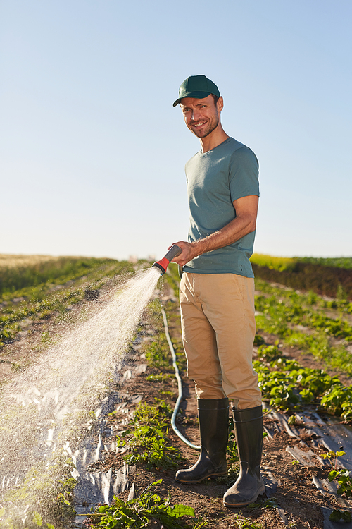 Vertical full length portrait of young male worker watering crops at vegetable plantation and smiling at camera while standing outdoors against blue sky
