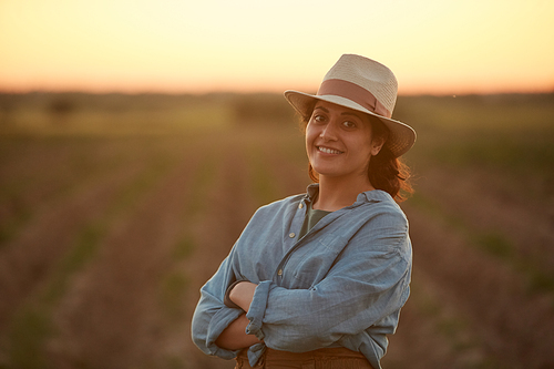 Waist up portrait of young female farmer posing confidently with arms crossed while standing in field at sunset and smiling at camera, copy space