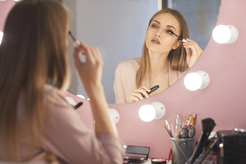 Reflection portrait of beautiful young woman applying mascara while doing makeup looking at vanity mirror, copy space