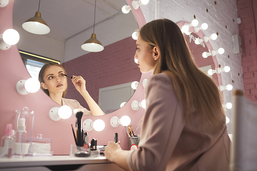Low angle portrait of beautiful young woman applying mascara while doing makeup looking at vanity mirror in pink interior, copy space