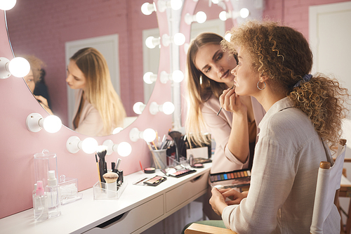 Side view portrait of female makeup artist styling fashion model by vanity mirror in pink dressing room interior, copy space