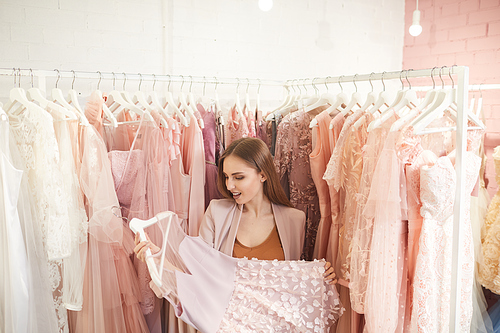 Waist up portrait of excited beautiful woman choosing glamorous dresses in clothing store, copy space