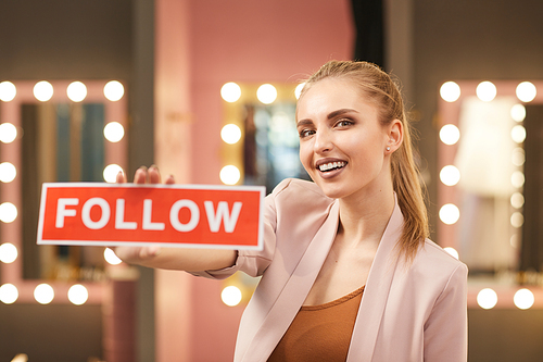 Portrait of beautiful young woman holding FOLLOW sign while posing in glamorous dressing room interior, copy space