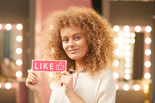 Portrait of modern curly-haired woman holding LIKE sign while posing in glamorous dressing room interior, copy space