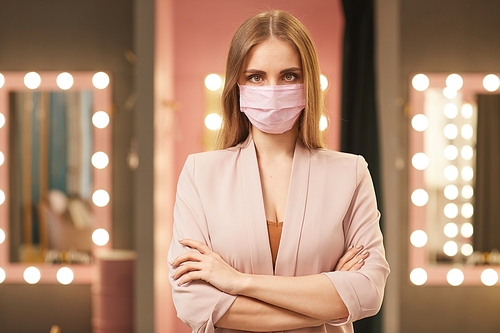 Waist up portrait of beautiful young woman wearing pink face mask standing with arms crossed while posing in dressing room interior, copy space