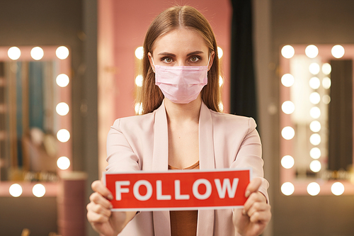 Waist up portrait of beautiful young woman wearing pink face mask and holding FOLLOW sign while standing in dressing room interior, copy space