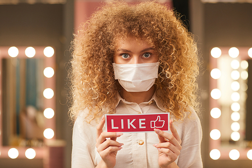 Waist up portrait of curly-haired young woman wearing face mask and holding Like sign while standing in dressing room interior, copy space