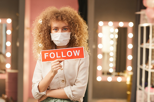 Waist up portrait of curly-haired young woman wearing face mask and holding Follow sign while posing in dressing room interior, copy space