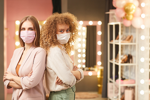 Waist up portrait of two modern young women wearing face masks standing back to back with arms crossed while posing in glamorous dressing room interior, copy space