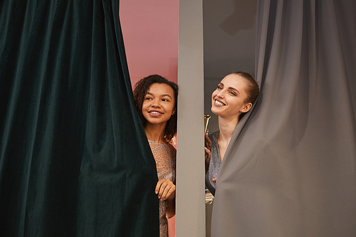 Portrait of two smiling young women peeking from dressing rooms while enjoying shopping in clothing store, copy space