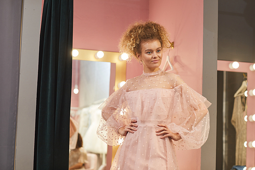 Portrait of modern young woman wearing pink dress posing in dressing room of clothing boutique, copy space
