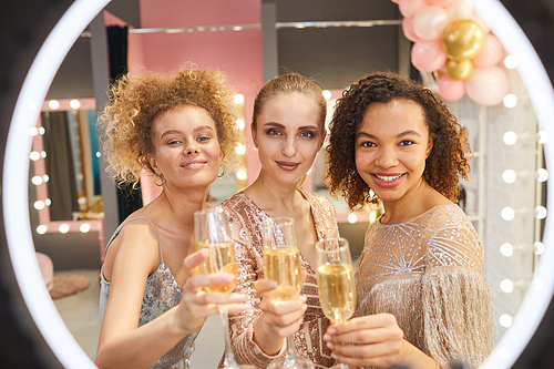 Multi-ethnic group of beautiful young women holding champagne glasses and smiling while  in dressing room interior, shot through ring light