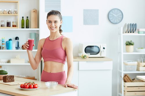 Waist up portrait of fit young woman holding smoothie while enjoying healthy lunch in kitchen interior and smiling at camera, copy space