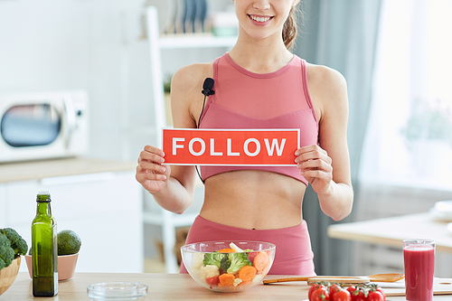 Mid section portrait of fit young woman holding FOLLOW sign and smiling at camera while cooking healthy food in kitchen, copy space