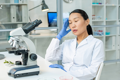 Young tired Asian female laboratory worker in gloves and whitecoat touching her head while sitting in front of microscope by workplace