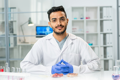 Young successful scientific researcher in whitecoat looking at you while sitting by workplace with samples of raw vegetable meat in petri dishes