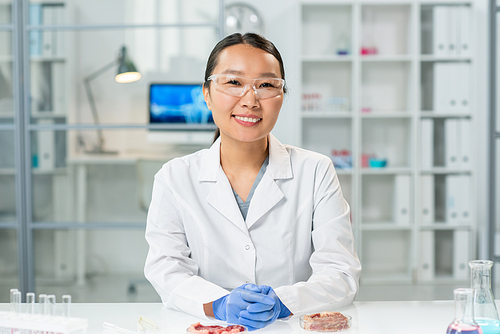 Happy young gloved Asian female in whitecoat and eyeglasses sitting by workplace with two samples of raw vegetable meat in petri dishes