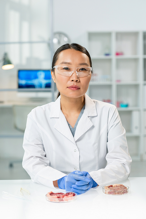 Young gloved Asian female researcher in whitecoat and eyeglasses sitting by workplace with two samples of raw vegetable meat in petri dishes