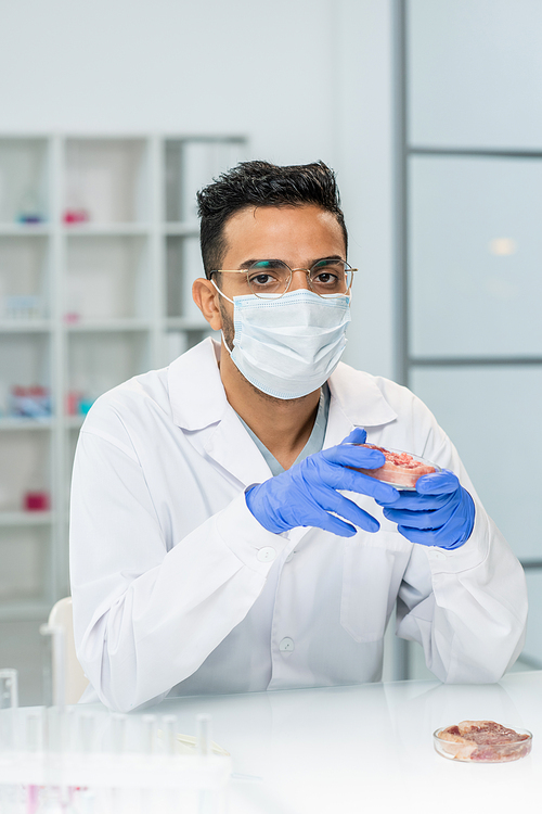 Young gloved male scientific researcher in whitecoat, mask and eyeglasses holding piece of raw vegetable meat in petri dish while working in lab