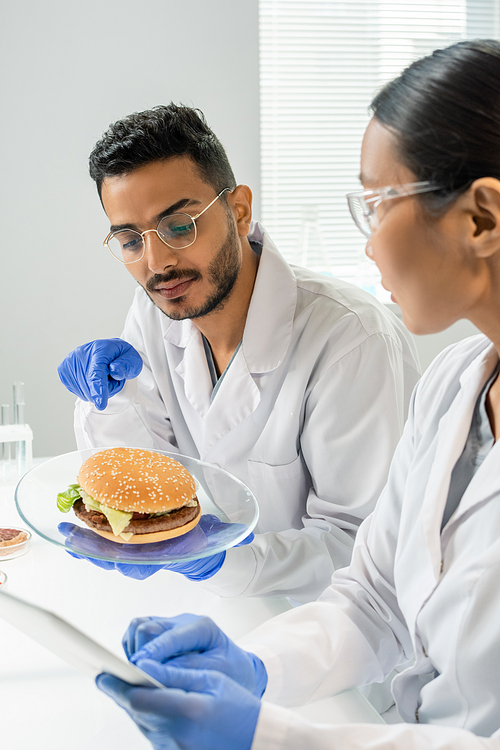 Gloved male worker of food quality control pointing at hamburger with vegetable meat while describing its characteristics to colleague