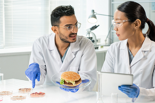 Gloved young man with hamburger on plate pointing at one of samples of raw vegetable meat while describing its characteristics to colleague