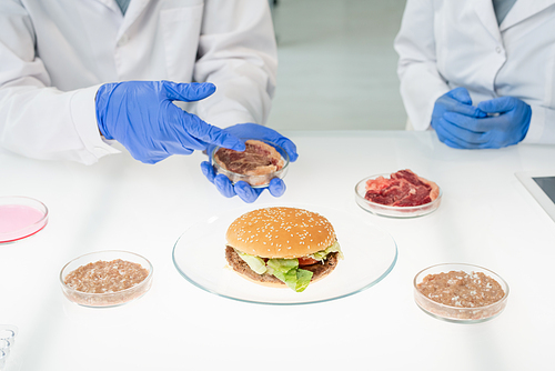 Gloved hands of food quality control worker holding petri dish with sample of raw vegetable meat over plate with hamburger and pointing at it