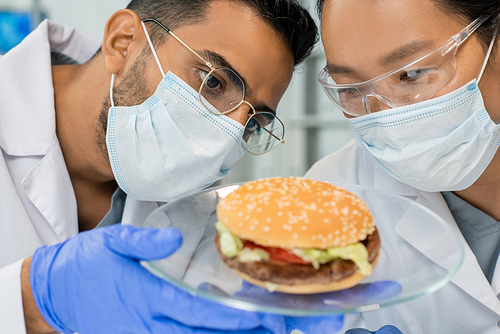 Two young intercultural lab workers in whitecoats, protective gloves, masks and eyeglasses looking at tasty hamburger containing vegetable meat