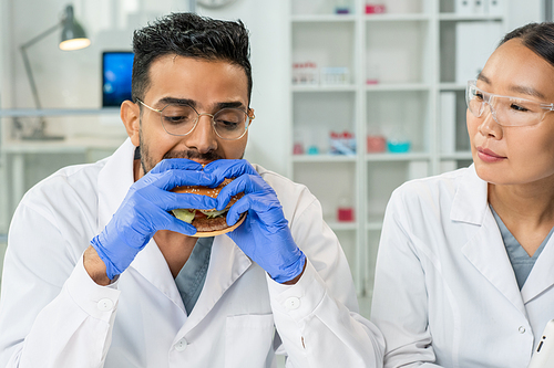Gloved young male worker of food control eating hamburger by workplace while checking quality and taste of its ingredients in laboratory