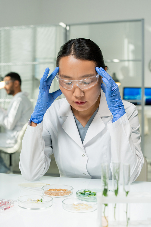 Young tired Asian female researcher in protective workwear touching her head and bending over desk while studying lab-grown sprouts