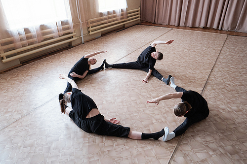Four young flexible dancers in avtivewear sitting with outstretched legs during training on the floor of modern dance studio