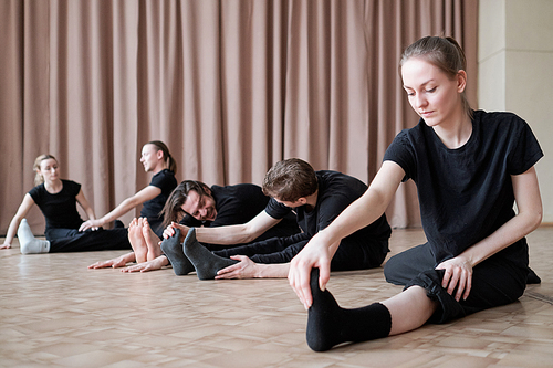 Fit young female dancer stretching one leg while sitting on the floor during training on background of other dancers exercising