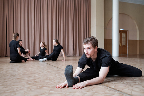 Young flexible man in black activewear sitting in twine position while training on the floor with group of dancers on background