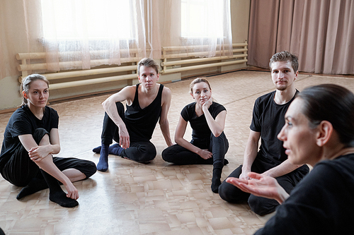 Group of young fit men and women in black activewear sitting on the floor and listening to their instructor of studio dancing