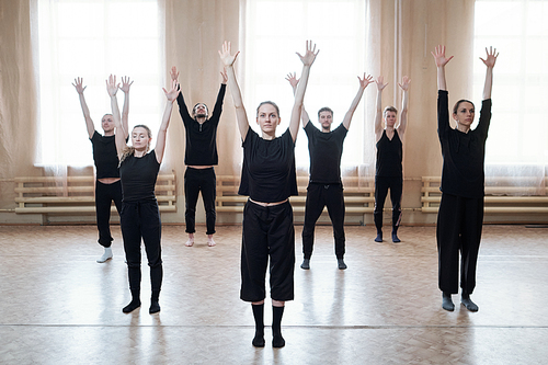 Group of young dancers in black activewear raising arms while standing on the floor against window in modern dance studio