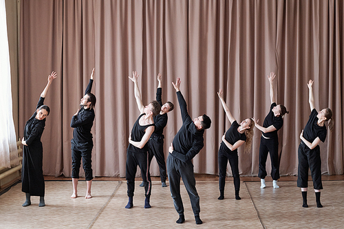 Five guys and three girls in black activewear stretching one arm upwards while standing on the floor during training in studio