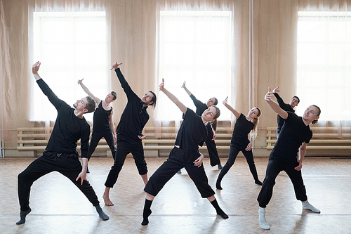 Group of young active dancers in black t-shirts and pants stretching one arm in front of themselves during dance exercise