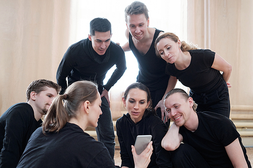 Group of young male and female dancers wearing black clothes spending time together watching something on smartphone