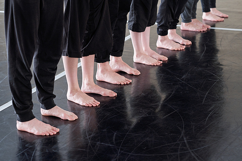 Row of legs of barefoot young dancers in black pants standing on the floor of dancing studio while exercising at lesson