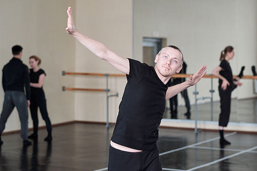 Young fit blond man in black activewear doing one of dance exercises while training in front of camera in studio of modern dancing