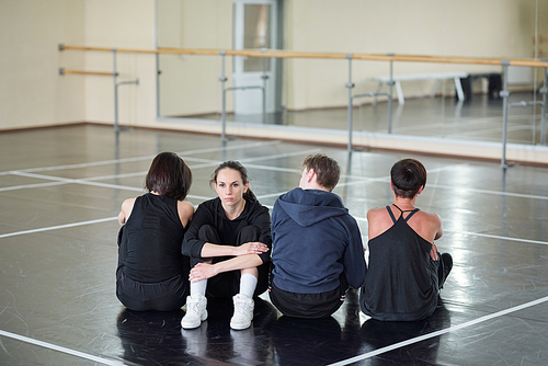 Young serious woman in costume  while sitting among three dancers on the floor of modern dance studio