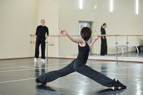Young fit and flexible woman in black activewear and sneakers doing stretching exercise while training in dance studio