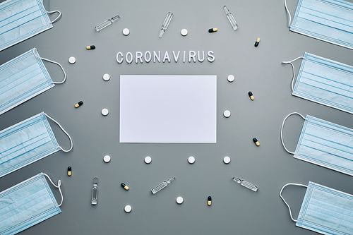 Above view composition of medical masks framing blank white paper with coronavirus word and medication laid out over grey background, copy space